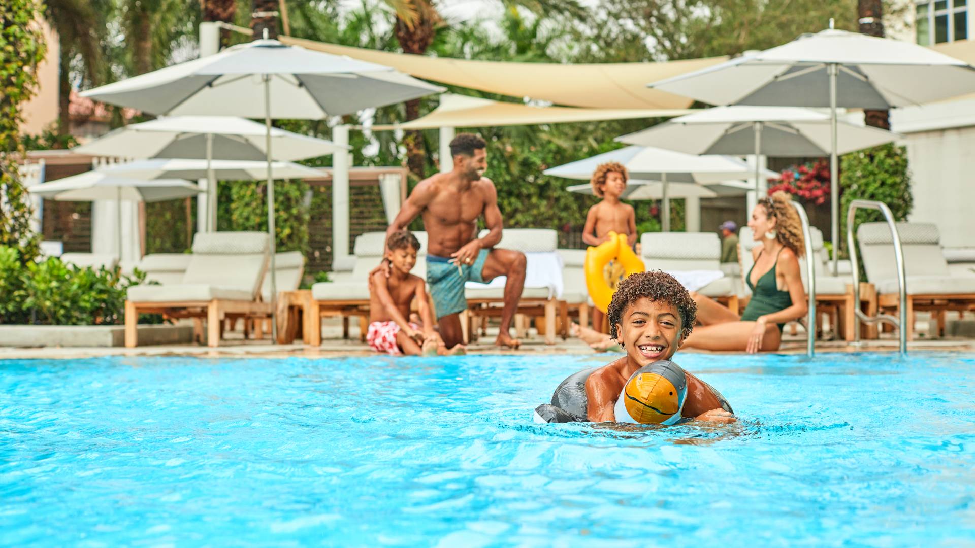 Family relaxing by the pool, with young child smiling as he floats in water