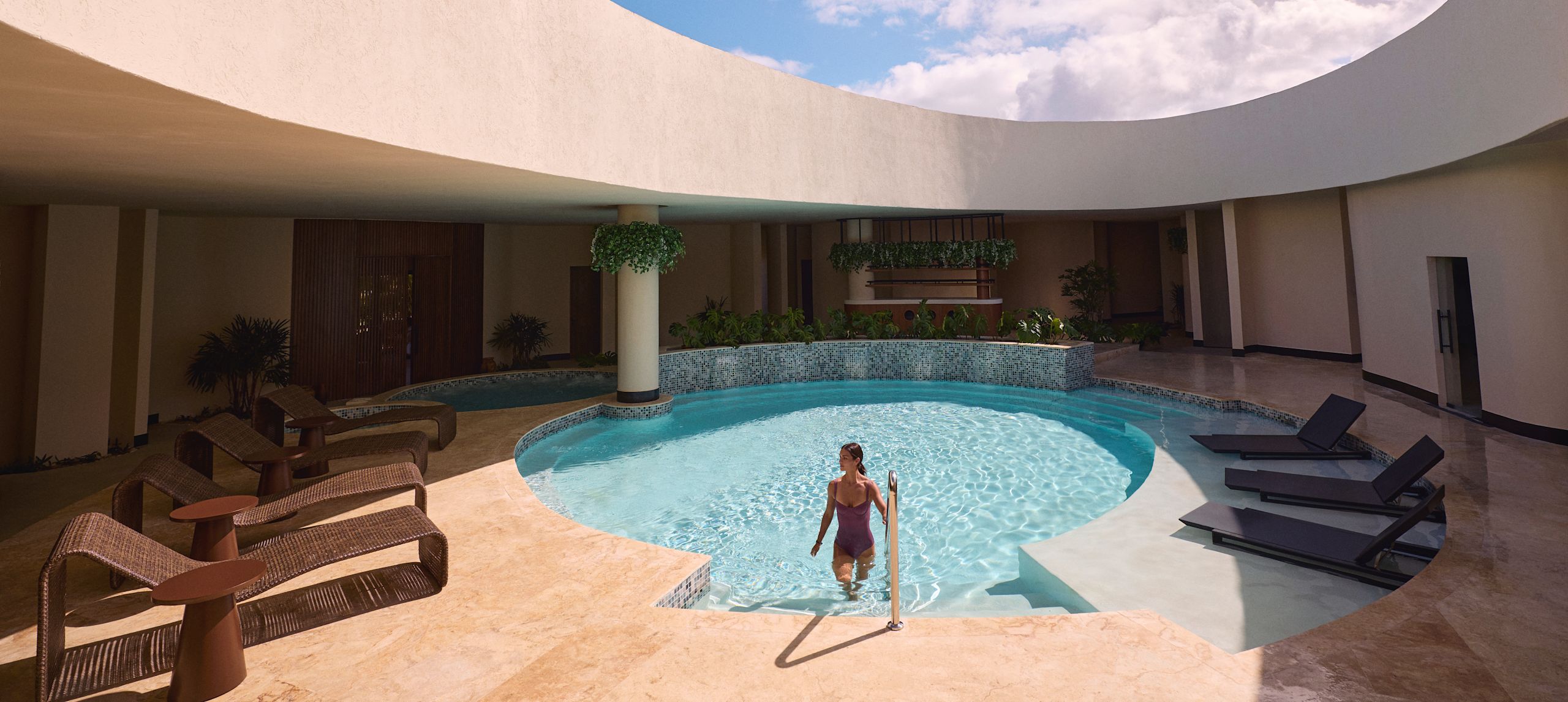 Person stepping out of a circular pool beneath an open skylight, surrounded by curved architecture, lounge chairs, and bright blue sky.