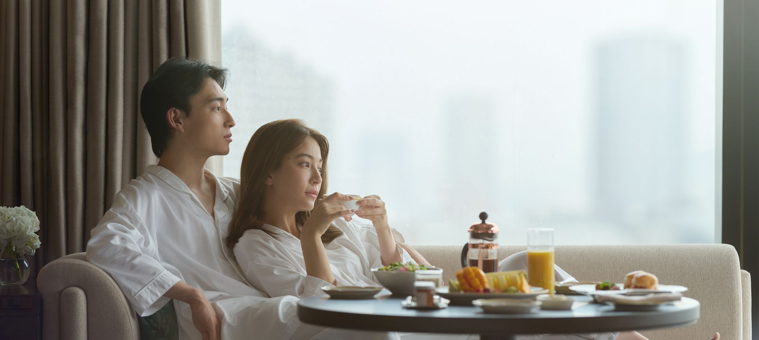 Pasangan mengenakan piama putih yang duduk-duduk di sofa di suite presidensial, dan sarapan tersedia di meja di depan mereka.-transisi sebelumnya