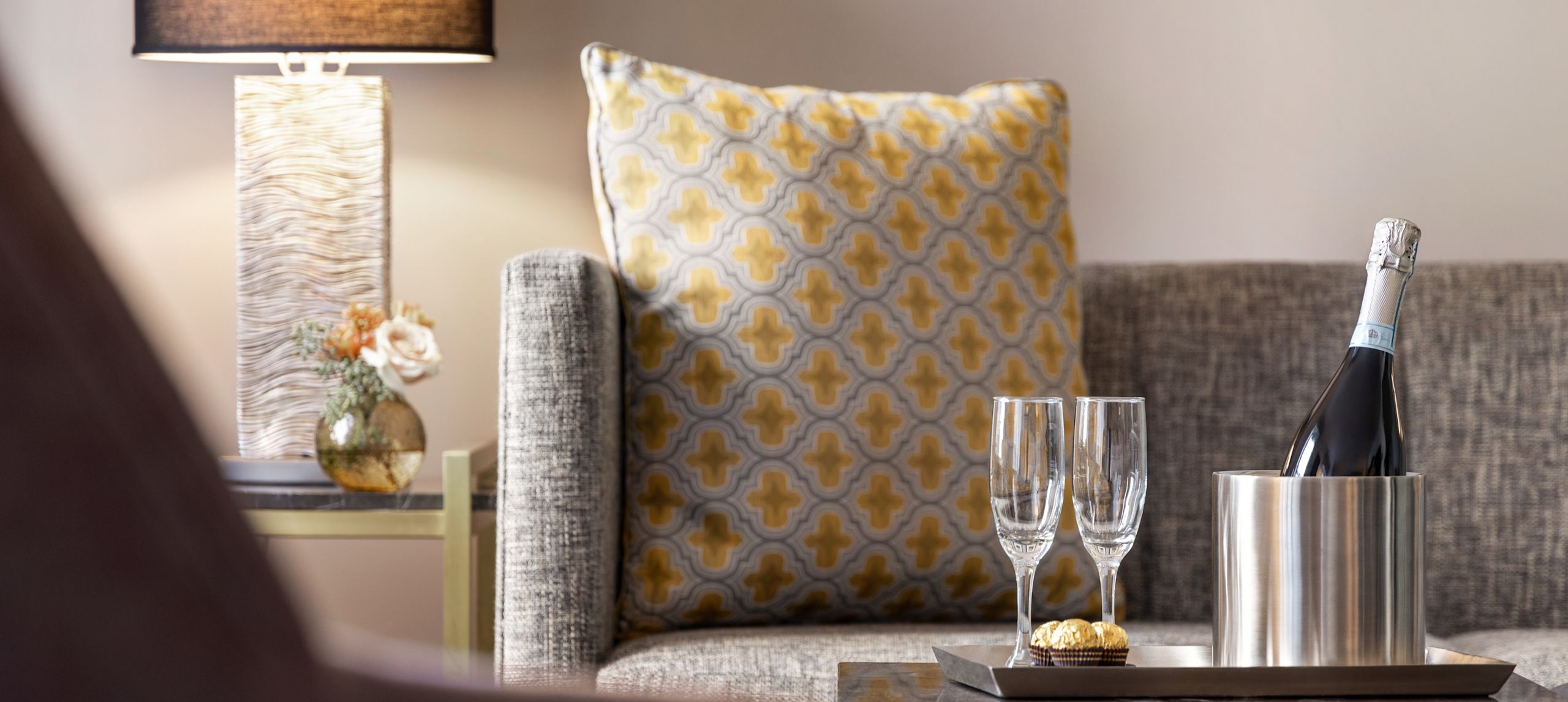 The end of the sofa, in the lounge area, with yellow, white, and gray decorative pillows and a tray with champagne and glasses on the table.