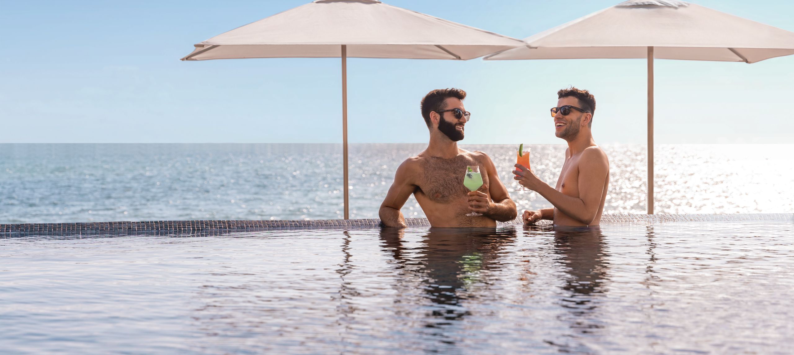 Two men stand in pool with drinks in hand