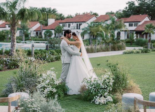 Bride and groom embrace in a garden with white floral arch and palm trees in the background.