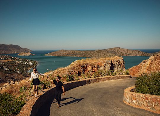 Couple walking on a curved pathway overlooking the sea and hills under a clear blue sky.