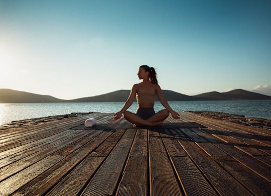 A woman practicing yoga on a wooden deck near a body of water, with hills in the background and clear blue sky.