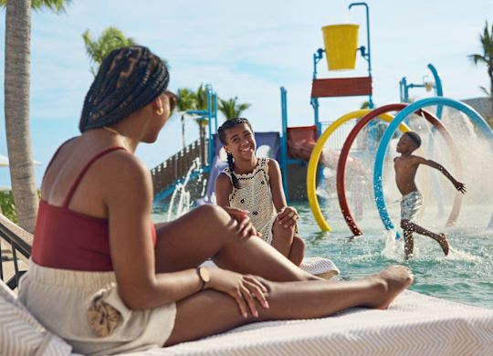 La mère et sa fille sont assises sur des chaises longues de la piscine et partagent un moment amusant pendant que le fils joue dans la Splash Zone de la piscine