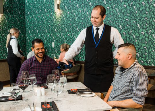 two men sitting at a dinner table with a server pouring wine
