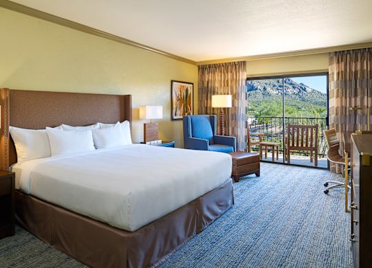 King Bed and Bedside Tables Facing TV and Work Desk with Wall Art and Reading Chair in Corner by Balcony with Mountain View