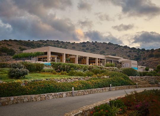 Hotel building exterior surrounded by lush vegetation on a hill at sunset, under a partly cloudy sky.