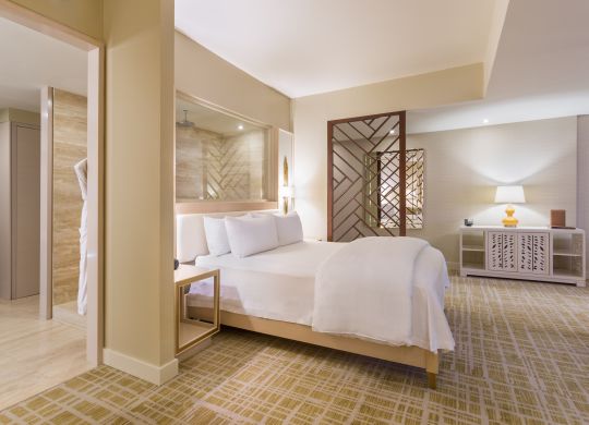 King suite bedroom with king bed, nightstands, lamps, and partial view of bathroom with vanity mirror, seat, and robes