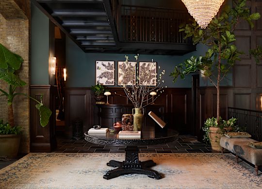 Hotel lobby with dark wood paneling, a chandelier, a central table with vases and books, and framed art on the wall.
