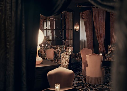 Luxurious dimly lit bar with velvet chairs and patterned carpet, dark curtains and walls, with candles on tables.