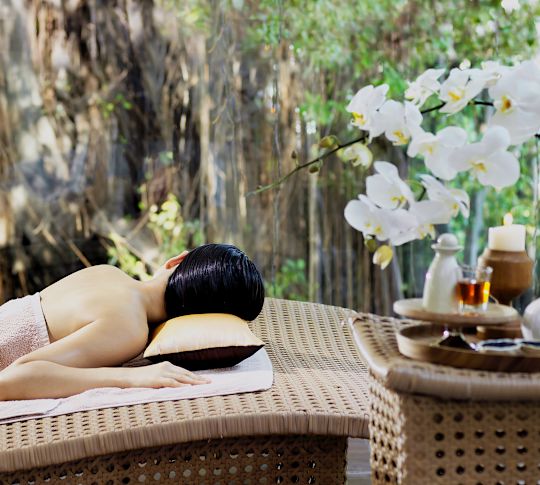 Una signora sdraiata su un lettino per massaggi è coperta da un asciugamano di spalle alla telecamera. In primo piano sono presenti oli per massaggi e fiori e sullo sfondo un tranquillo giardino.