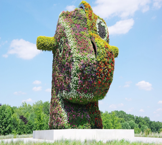 Värikkäiden kukkien peitossa oleva veistos seisoo valkoisella jalustalla