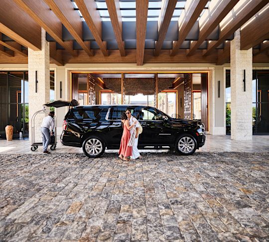 Zwei Frauen umarmen sich mit einem großen schwarzen Auto. Mitarbeiter kümmert sich um Gepäck