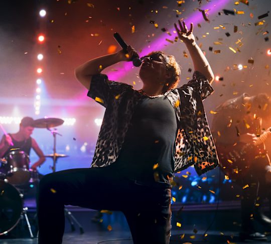 Artista cantando en el escenario con banda detrás y confeti lloviendo con dardo