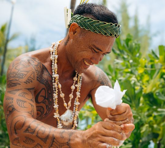 Polynesian man wearing traditional garments and accessories, is preparing fresh coconut milk 