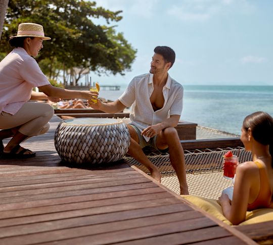 Hombre y mujer relajándose junto al mar mientras un miembro del personal entrega bebidas