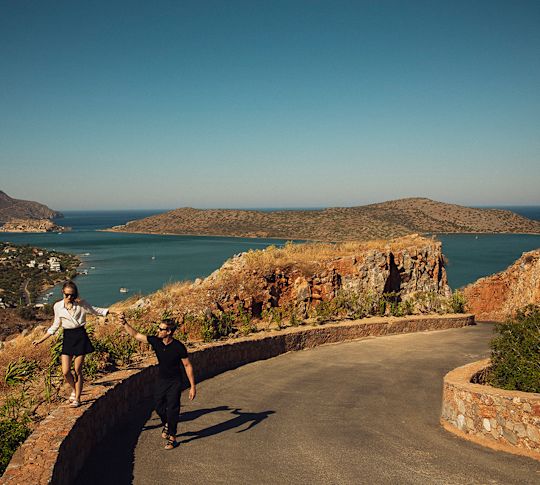 Pár kráčí po zakřivené cestě s výhledem na moře a kopce pod jasnou modrou oblohou.