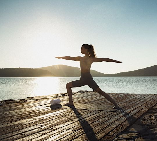 Woman practicing yoga on a wooden deck by the sea during sunrise.