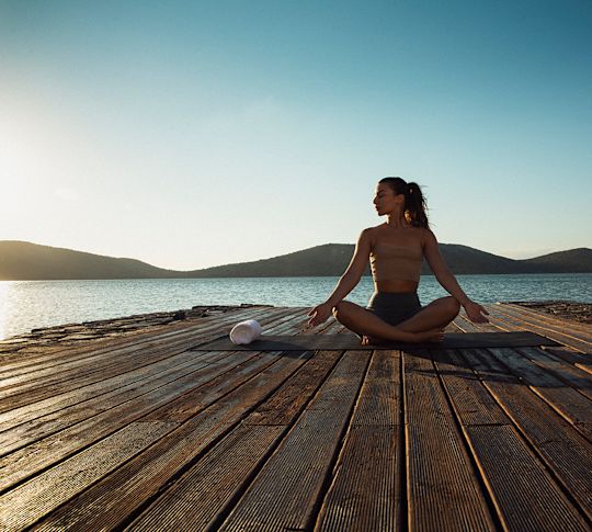 Žena procvičující jógu na dřevěném palubě poblíž vodní hladiny, s kopci v pozadí a průzračně modrou oblohou.