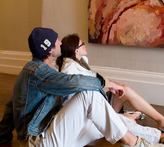 A young woman and men in fashionable leisure clothing are sitting on the floor of an art gallery looking at a large painting
