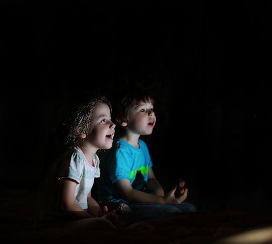 Two young kids sitting in darkened room, illuminated by a screen.
