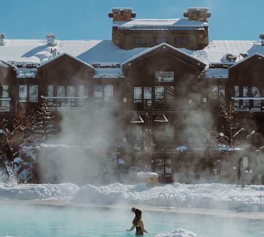 A Woman in an Outdoor Hot Pool on a Snowy Day, and Hotel Exterior Showing in Background