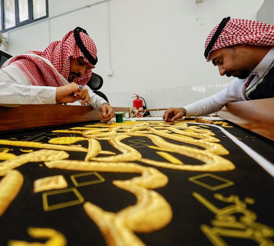 Two Arabic men with checkered head coverings, sewing the gold letterings on a black tapestry.