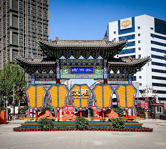 Poartă chinezească festivă cu felinare galbene suspendate și detalii complexe, încadrate de ascensiuni urbane înalte.