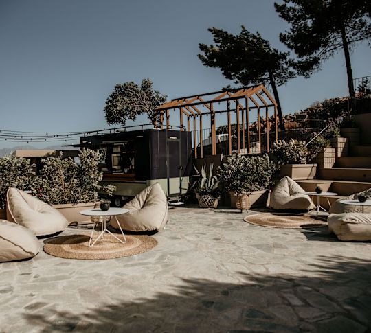 Amphiteatro Büyük bej fasulye çantaları, yuvarlak kurabiye paspasları, taş döşemeler bulunan, yeşil çalılar ve ağaçlarla çevrili açık oturma alanı.