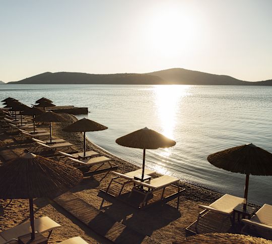 Pláž s řadami lehátek a deštníků při západu slunce, s horami v dálce.