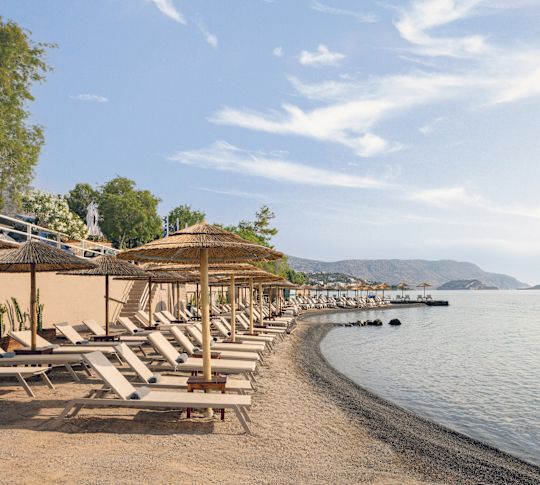 Coastal view of a serene beach with lounge chairs and straw umbrellas, overlooking the clear blue sea and surrounding hills.