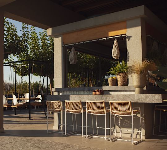 Fitil tabureleri, gri bir tezgâh, saksı bitkileri ve ağaçlar ve masalarla çevrili asılı Fitil ışıkları bulunan açık bar tezgahı.