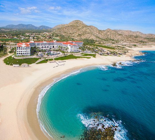 Aerial view of Hilton Los Cabos Beach & Golf Resort. Beach and coastline in the foreground. Hotel and mountains in the background.