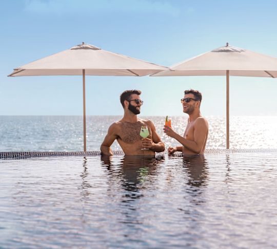 Two men stand in pool with drinks in hand