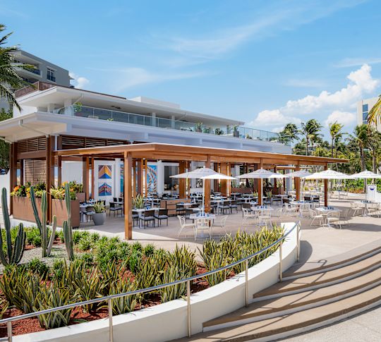 Outdoor restaurant patio with dining tables and umbrellas, surrounded by palm trees, planters, and curved steps.