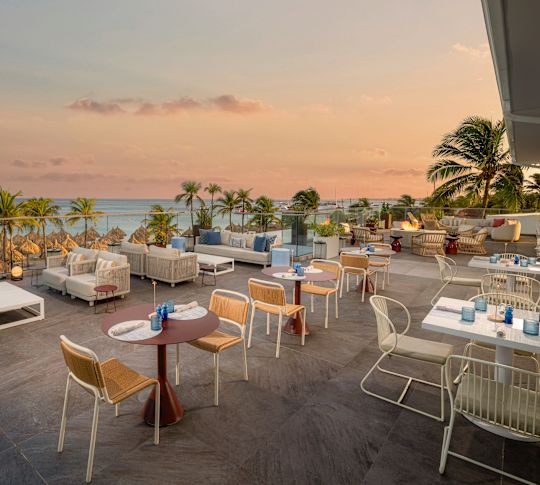 Outdoor restaurant terrace with dining tables and lounge seating overlooking palm trees and the ocean at sunset.