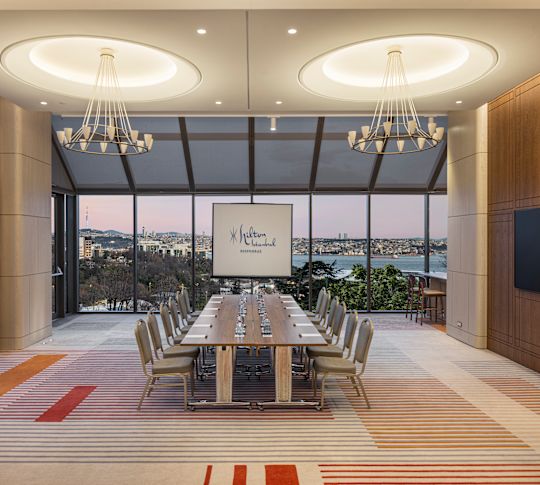 Meeting room with a long conference table, chairs, a presentation screen, and floor-to-ceiling windows overlooking the city and water