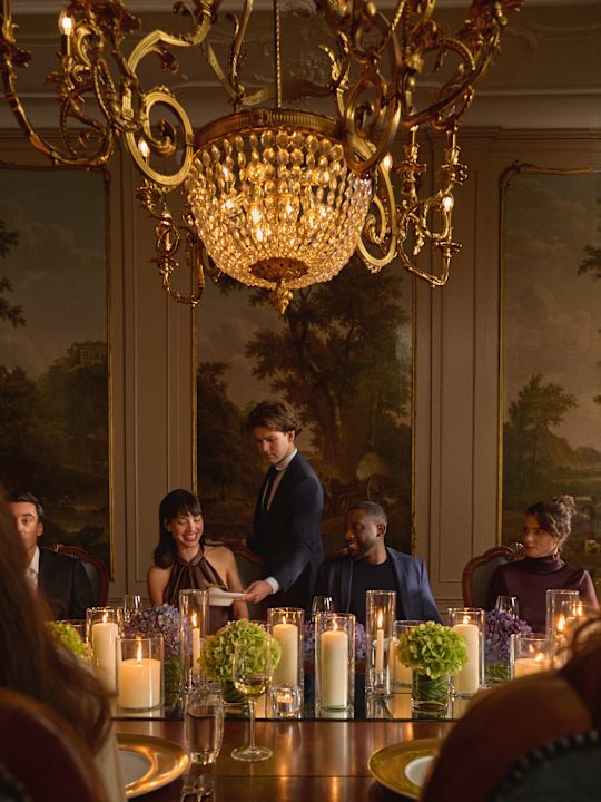 Formal dinner setting in an opulent dining room with a large crystal chandelier, ornate wall panels, and a long table decorated with candles and green floral arrangements, with several people seated and a server standing.