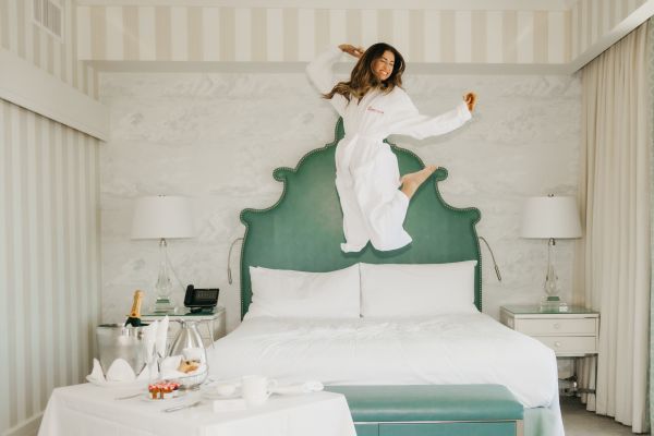 Woman jumping on bed, with room service items in foreground