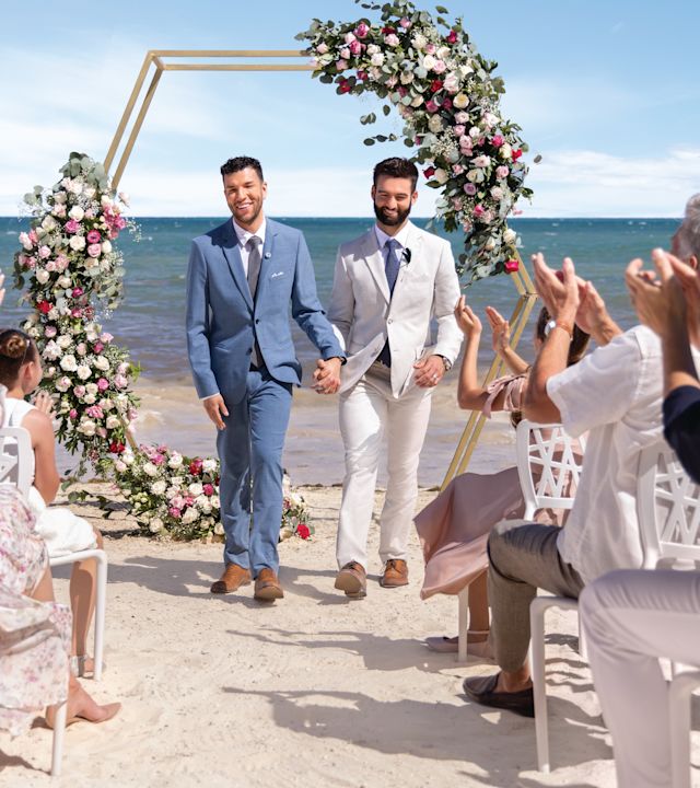 Two men in suits walk down the aisle on the beach while guests clap
