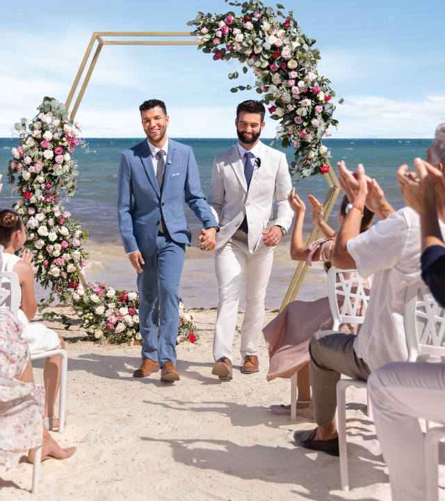 Two men in suits walk down the aisle on the beach while guests clap