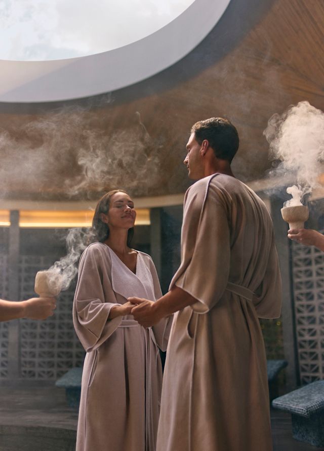 O femeie și un bărbat țin de mână în timp ce stau în centrul spa, iar membrii echipei țin boluri cu tămâie pentru fumat