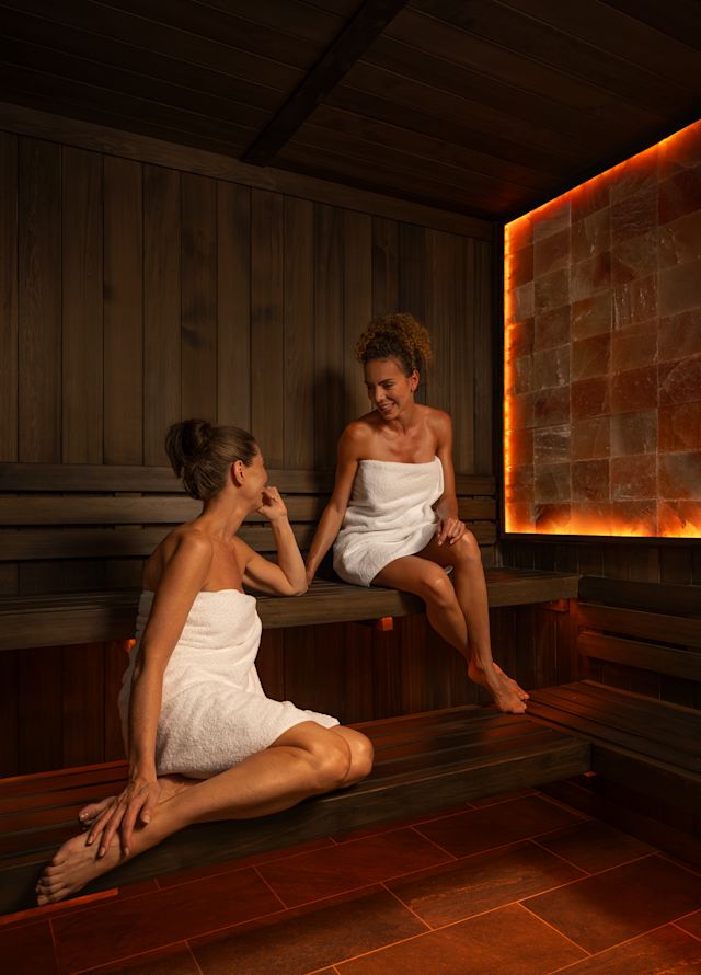 Ahşap ve sıcak bir kaya duvarından yapılmış saunada oturan havlulu iki kadın.