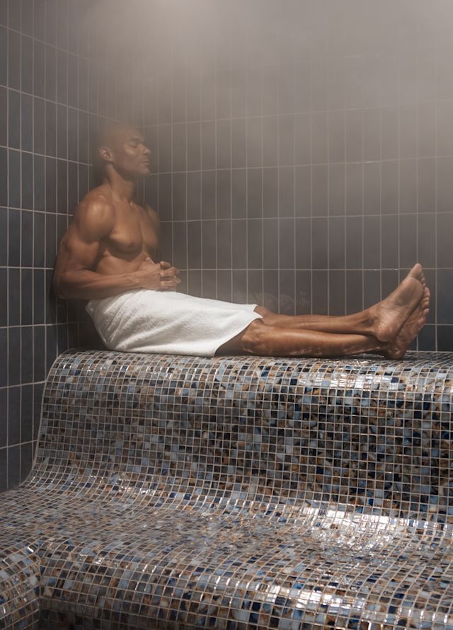 Buhar odasında oturan, havlu giyen ve bacakları açık ve çapraz olan adam.