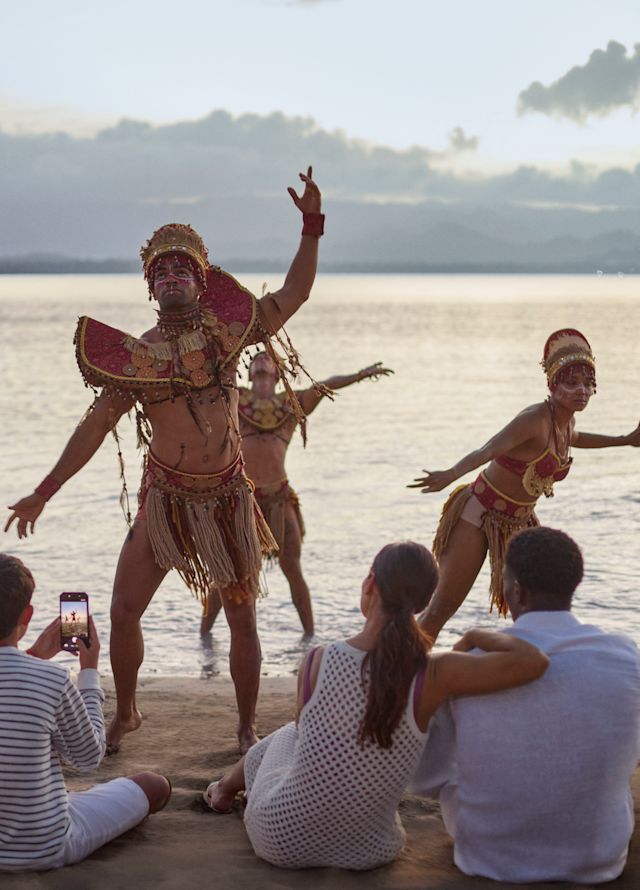Dancers performing a traditional beachside dance at sunset while a seated group watches, with the ocean and distant hills in the background.