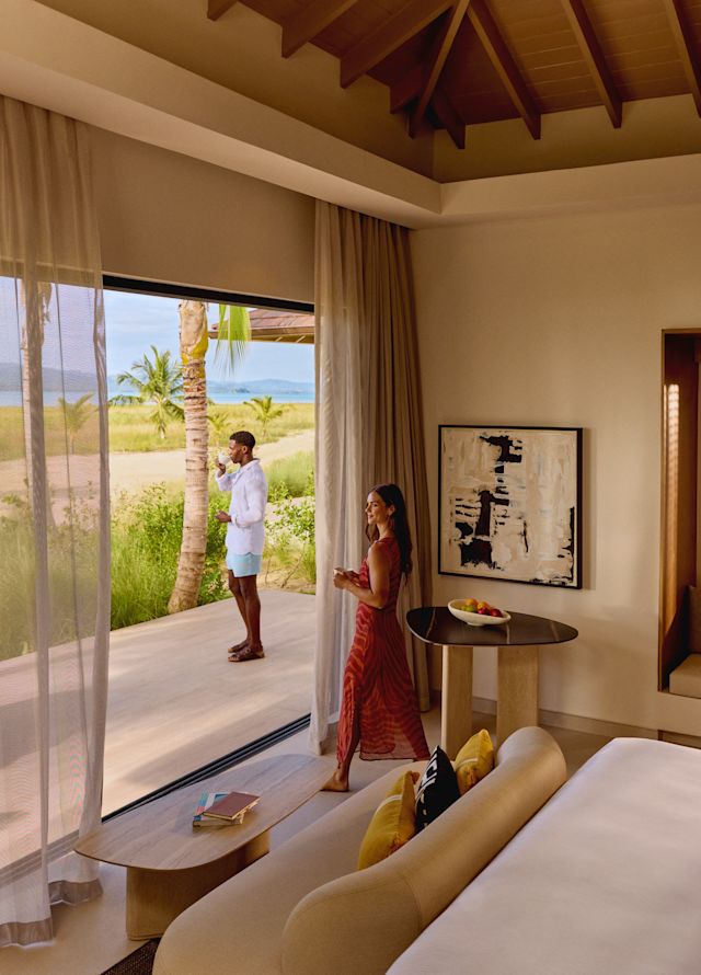 Two people in a sunlit bedroom suite, one standing on the deck looking toward the landscape while the other enjoys a drink near the open sliding doors, with views of palm trees and distant water.