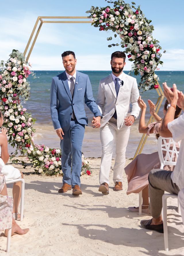 Two men in suits walk down the aisle on the beach while guests clap
