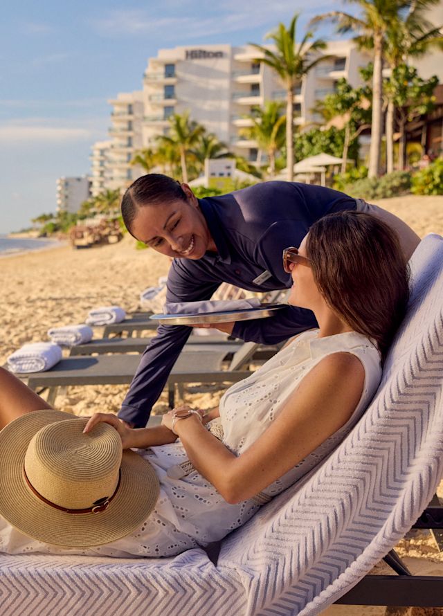 A woman is relaxing on a sun lounger on the beach and talking to a staff member while a couple are jogging past