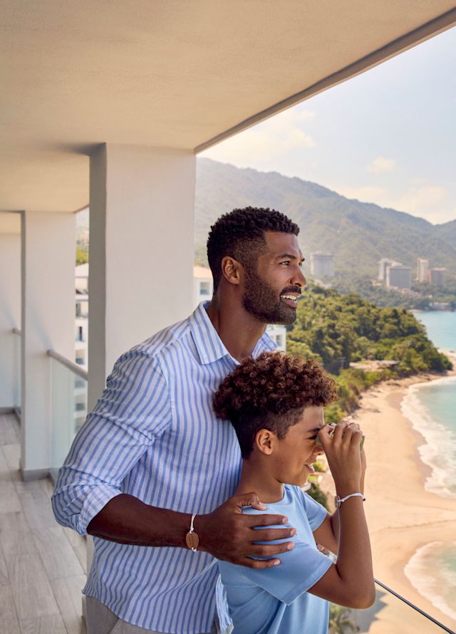 Father and son standing on suite balcony looking out to the sea with binoculars